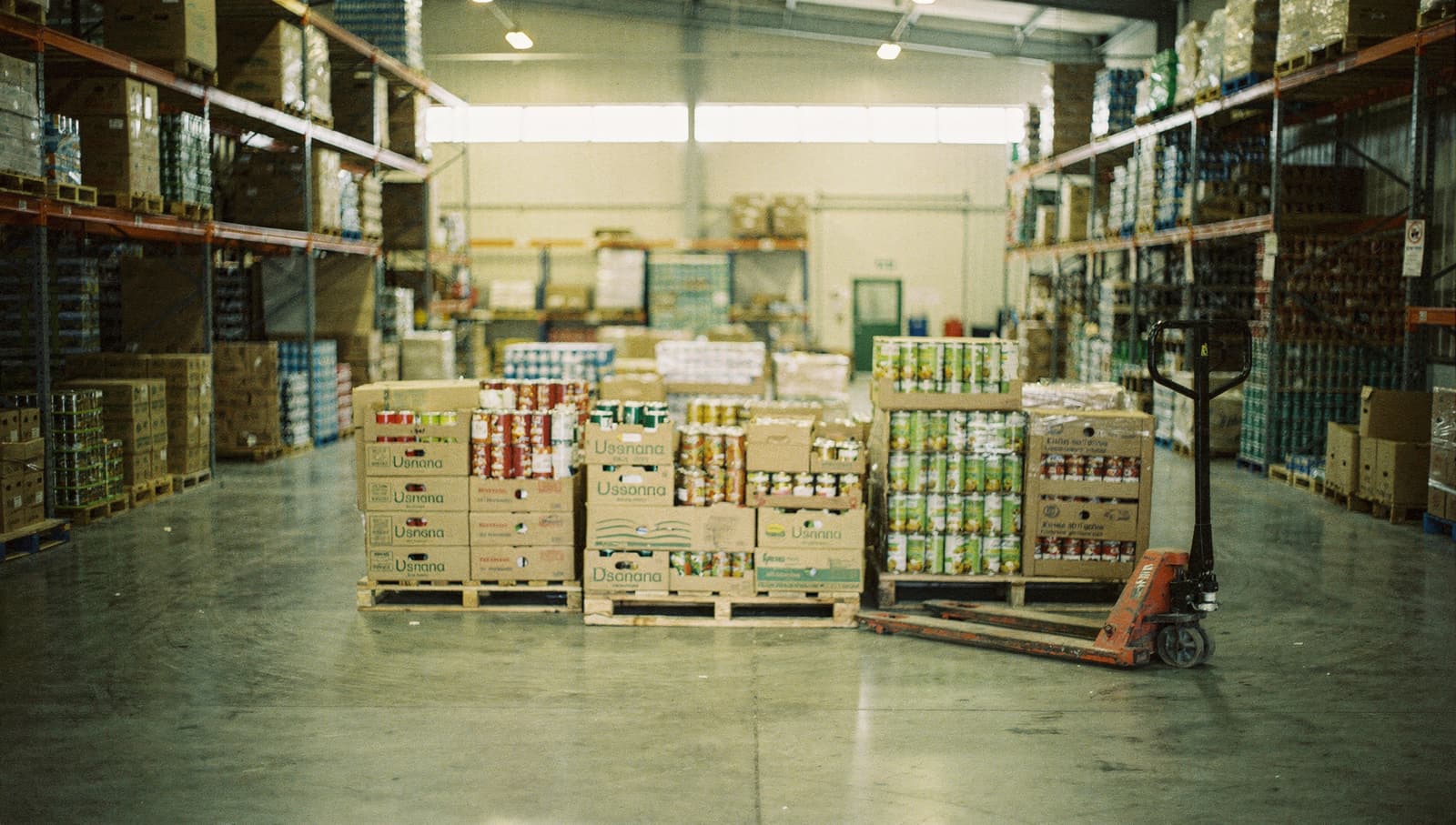 A food bank warehouse: pallets stacked with boxes and trays of tinned food, tall racking down both sides, a pallet jack parked to one side.